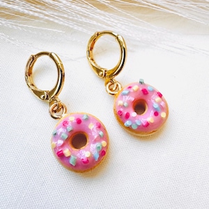 Puede incluir: Un par de pendientes de aro dorados, cada uno con un pequeño dije de donut rosa. Los donuts están decorados con chispas de colores. Los pendientes se exhiben sobre una superficie blanca.