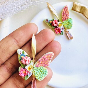 May include: A pair of gold butterfly earrings with pink, white, and green wings. The wings are made of polymer clay and have a textured look. The earrings have a pink crystal dangle.
