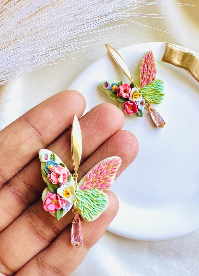 May include: A pair of gold butterfly earrings with pink, white, and green wings. The wings are made of polymer clay and have a textured look. The earrings have a pink crystal dangle.