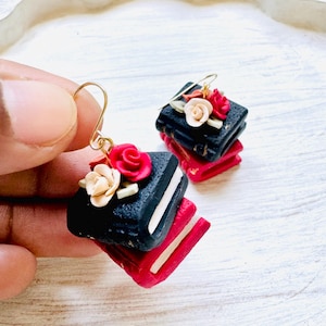 May include: A pair of dangle earrings featuring a stack of three miniature books with red and black covers. The top book has a small cluster of red, white, and cream colored flowers.