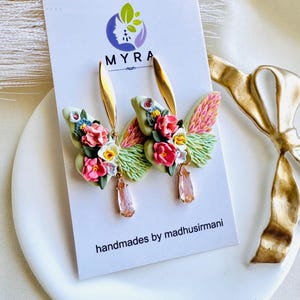 May include: A pair of floral butterfly earrings with pink, white, and yellow flowers. The earrings are made of polymer clay and have a gold-tone finish. The earrings are hanging from a gold-tone hook.