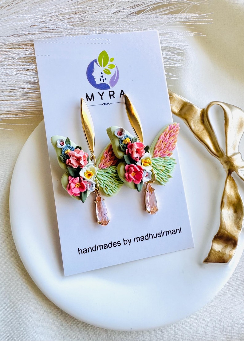 May include: A pair of floral butterfly earrings with pink, white, and yellow flowers. The earrings are made of polymer clay and have a gold-tone finish. The earrings are hanging from a gold-tone hook.