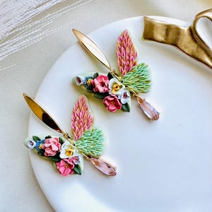 May include: A pair of floral butterfly earrings with pink, green, and white accents. The earrings feature a delicate crystal drop and gold-tone hardware.