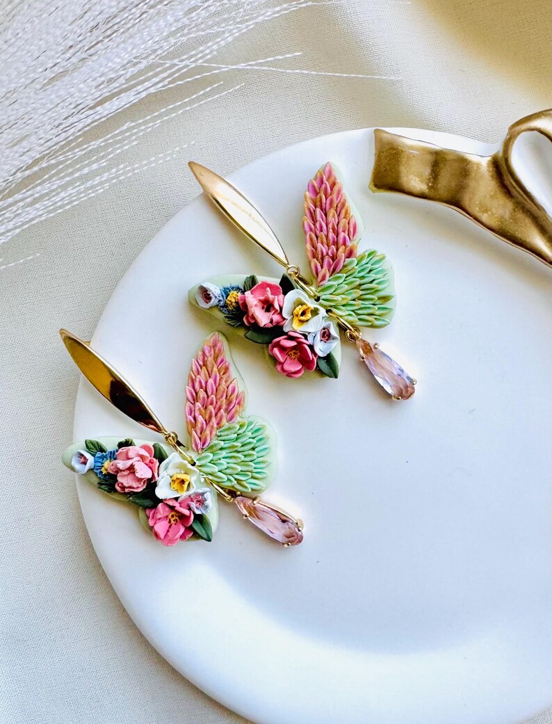 May include: A pair of floral butterfly earrings with pink, green, and white accents. The earrings feature a delicate crystal drop and gold-tone hardware.