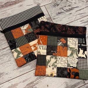 May include: Two square fabric zipper pouches featuring a patchwork design of various Halloween-themed patterns including ghosts, pumpkins, black cats, spiderwebs, and houndstooth in shades of orange, black, cream, and green with black zippers.