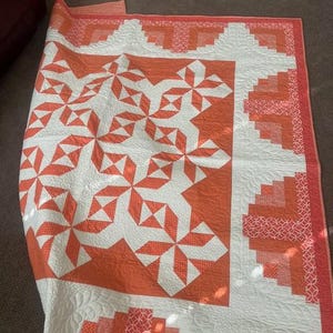 May include: A white and orange quilt with a geometric pattern. The quilt is folded in half and lying on a brown carpet.