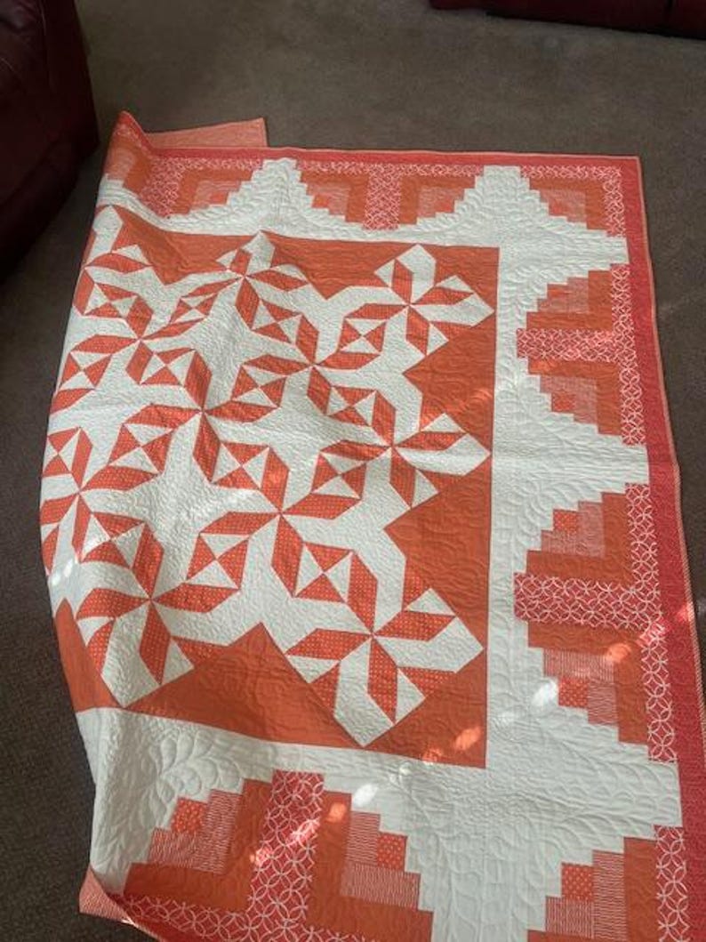 May include: A white and orange quilt with a geometric pattern. The quilt is folded in half and lying on a brown carpet.