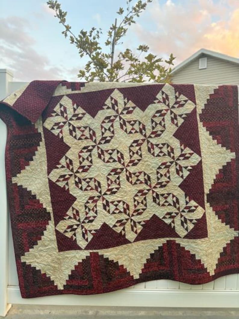May include: A quilt with a burgundy and cream color scheme. The quilt features a geometric pattern with a central star design. The quilt is hanging on a white fence.