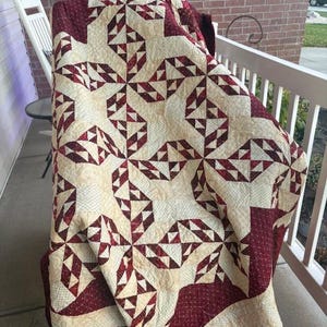 May include: A red and cream quilt with a geometric pattern of triangles. The quilt is folded and draped over a chair on a porch.
