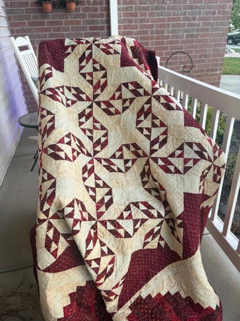 May include: A red and cream quilt with a geometric pattern of triangles. The quilt is folded and draped over a chair on a porch.