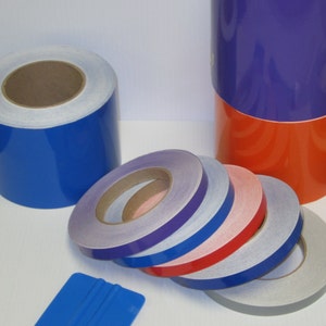 May include: Assortment of reflective tape rolls in various colors, including blue, purple, red, and gray. The rolls are stacked on a white surface. A blue squeegee is also visible in the image.