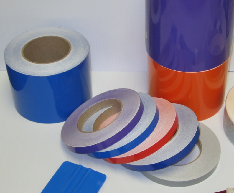 May include: Assortment of reflective tape rolls in various colors, including blue, purple, red, and gray. The rolls are stacked on a white surface. A blue squeegee is also visible in the image.