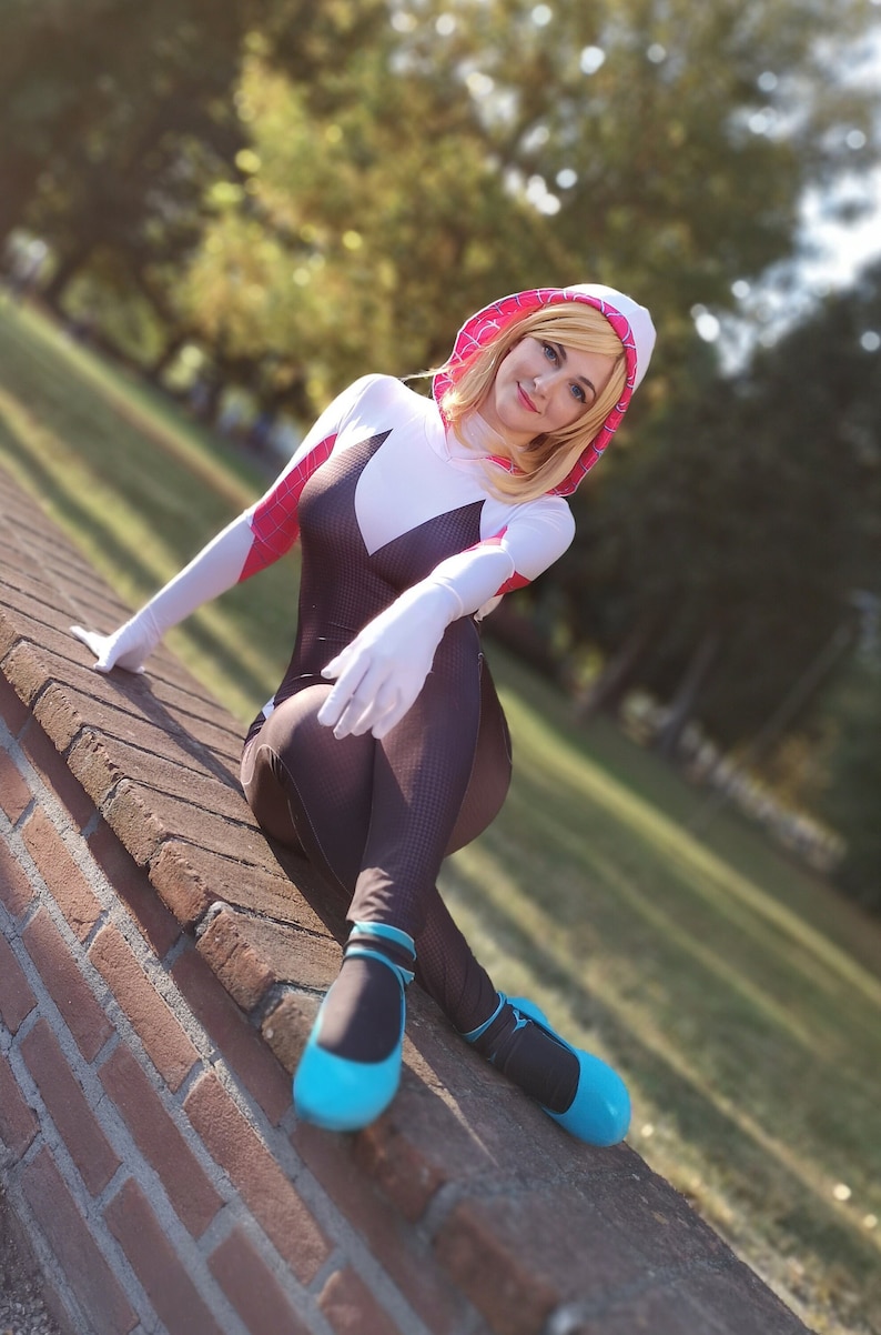 May include: A woman in a white and black spider-man costume with a pink hood sits on a brick wall. She is wearing blue shoes and has blonde hair.