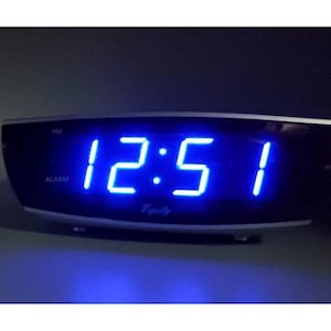 Könnte beinhalten: Ein digitaler Wecker mit gewölbter schwarzer Front und silbernem Rand. Das Display zeigt leuchtend blaue LED-Ziffern mit der Uhrzeit 12:51. Unter den Ziffern steht in weißer Schreibschrift das Wort Equity, sowie PM- und ALARM-Anzeigen.