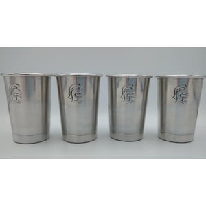 May include: A set of four silver-toned metal tumblers arranged in a row. Each cylindrical cup features a debossed graphic of a Spartan helmet profile on the front surface. The cups have a smooth, polished finish and a simple, straight-sided design.