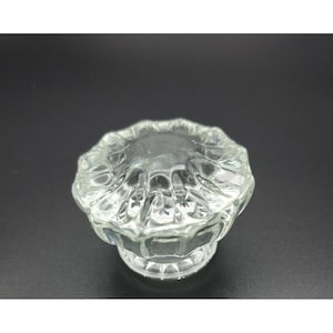 May include: A clear glass cabinet knob featuring a circular, multi-faceted design with a scalloped edge. The top surface has a series of radiating indentations that create a star-like pattern, while the base is cylindrical and smooth.