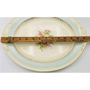 May include: An oval ceramic platter with a cream center, light blue rim, and gold filigree border. A floral bouquet is painted in the center. A wooden ruler across the platter shows a width of approximately 13 inches, with text WAGGONER'S SEED STORE.