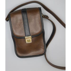 May include: A vertical rectangular crossbody bag in brown with black trim and a central black vertical stripe. The bag features a fold-over flap secured by a gold-tone metal clasp and includes a long, adjustable brown shoulder strap.