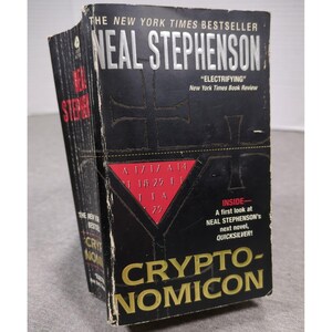 Pode incluir: Um livro de bolso intitulado Cryptonomicon de Neal Stephenson com uma capa preta apresentando um triângulo vermelho contendo números e letras, texto prateado no topo e texto dourado na parte inferior. A capa inclui um selo de best-seller.