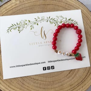 Peut inclure: Bracelet rouge en perles avec un charme en or en forme de fraise et le nom "AVA" écrit en lettres dorées. Le bracelet est sur une carte blanche avec le texte "Little Sparkles Boutique" et une adresse de site web.
