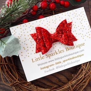 May include: A red glitter hair bow on a white card with gold and silver polka dots. The card has the text "Little Sparkles Boutique" and contact information for the shop's website, Instagram, and Facebook.