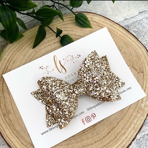 May include: Gold glitter hair bow with a sparkly finish. The bow is displayed on a white card with the text "Little Sparkles Boutique" and social media icons.