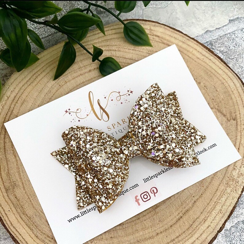 May include: Gold glitter hair bow with a sparkly finish. The bow is displayed on a white card with the text "Little Sparkles Boutique" and social media icons.