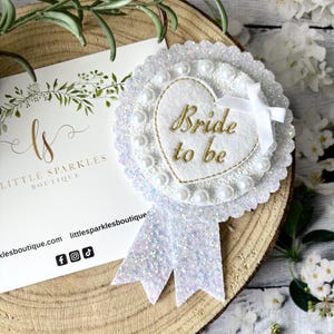 Könnte beinhalten: Weißer Braut-zu-sein-Schärpe mit einem weißen Herzen und Perlenbesatz. Die Schärpe ist mit weißem Glitzer verziert und hat ein weißes Band.