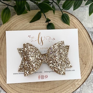 May include: A gold glitter hair bow with a white glitter center. The bow is on a white card with the text "Little Sparkle Boutique" and social media icons.