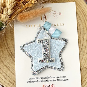 May include: A light blue glitter star with a white number one in the centre. The star is on a white card with the text "Little Sparkles Boutique" and a website address.
