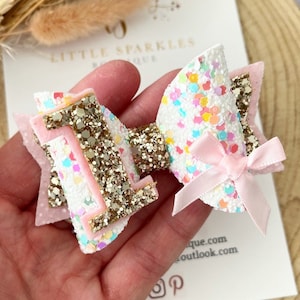 Puede incluir: Un lazo de brillo rosa y blanco con un número uno de brillo dorado. El lazo tiene una cinta de satén rosa en el centro. El lazo es perfecto para una fiesta de primer cumpleaños.