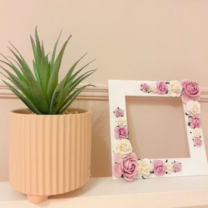 Op de afbeelding: Wit fotolijstje versierd met roze en witte bloemen. Het lijstje staat op een witte plank met een potplant links.