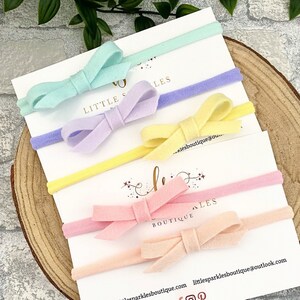 Pode incluir: Cinco laços de feltro em cores pastel com faixas elásticas combinando. Os laços estão dispostos em uma pilha, com a cor mais clara no topo e a cor mais escura na parte inferior. As cores são azul claro, lavanda, amarelo, rosa e pêssego.