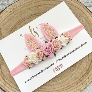 Op de afbeelding: Een roze haarband met konijnenoren en bloemenaccenten. De oren zijn roze met glitter, en de bloemen zijn in roze en witte tinten. De tekst "Little Sparkles Boutique" is zichtbaar.