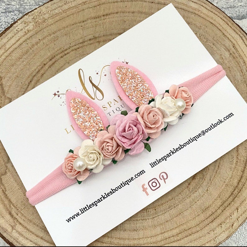 Op de afbeelding: Een roze haarband met konijnenoren en bloemenaccenten. De oren zijn roze met glitter, en de bloemen zijn in roze en witte tinten. De tekst "Little Sparkles Boutique" is zichtbaar.