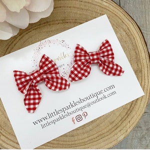 May include: Two red and white gingham hair bows. The bows are tied with a simple knot and have a slightly ruffled texture. The bows are displayed on a white card with the text "www.littlesparklesboutique.com littlesparklesboutique@outlook.com f  p".