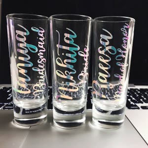 Puede incluir: Tres vasos de chupito de vidrio transparente con letras iridiscentes. Los vasos est&aacute;n etiquetados como "Bridesmaid", "Bride" y "Maid of Honor".
