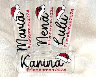 May include: Four clear shot glasses with black lettering and a red Santa hat design. The glasses are labeled "Mania", "Mena", "Lula", and "Karina" with the text "Friendsmas 2024" underneath each name.