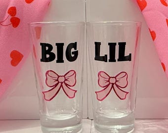 May include: Two clear glass shot glasses with pink bows and the words "BIG" and "LIL" printed on them.