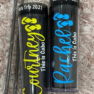 Peut inclure: Deux gobelets en plastique noir et transparent avec couvercles et pailles. Les gobelets portent le texte "Girls Trip 2021" en haut et les noms "Courtney" et "Rachel" en lettres jaunes et bleues. Le texte "This is Cabo" est &eacute;crit sous les noms.