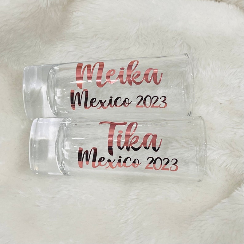 Puede incluir: Dos vasos de chupito de vidrio transparente con letras doradas. El vaso superior dice "Meika M&eacute;xico 2023" y el vaso inferior dice "Tika M&eacute;xico 2023".