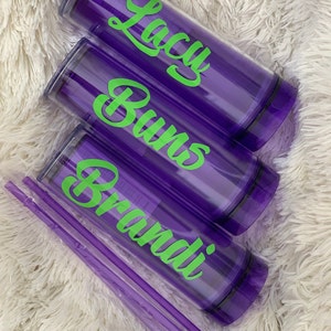 Peut inclure: Trois gobelets violets avec couvercles et pailles. Les gobelets portent des inscriptions vertes qui disent "Brandi", "Burns" et "Lacy".