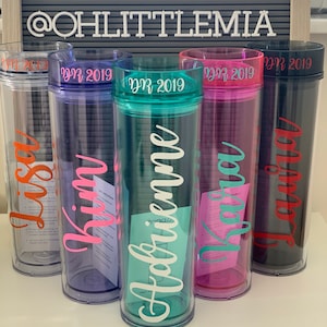 Peut inclure: Cinq gobelets en plastique transparent avec des couvercles de couleurs diff&eacute;rentes et des noms personnalis&eacute;s. Les noms sont "Alyssa", "Kim", "Gabrielle", "Kayla" et "Laurie". Chaque gobelet a un autocollant rose avec la date "MR 2019" dessus.