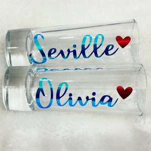 Puede incluir: Dos vasos de chupito de vidrio transparente con letras de vinilo azul y rojo. El vaso de arriba dice "Seville" con un coraz&oacute;n rojo. El vaso de abajo dice "Olivia" con un coraz&oacute;n rojo.
