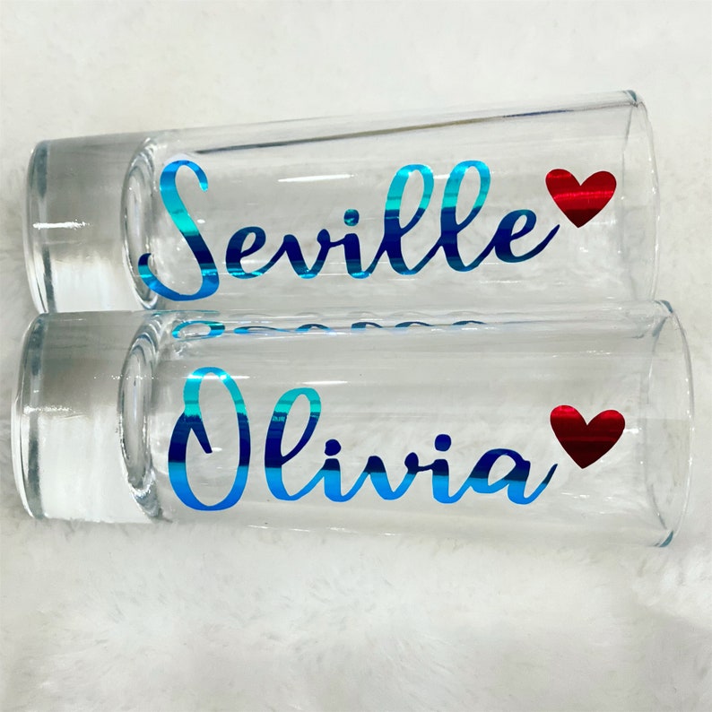 Puede incluir: Dos vasos de chupito de vidrio transparente con letras de vinilo azul y rojo. El vaso de arriba dice "Seville" con un coraz&oacute;n rojo. El vaso de abajo dice "Olivia" con un coraz&oacute;n rojo.