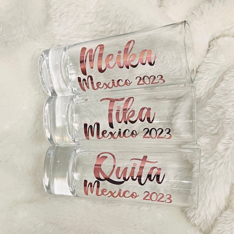 Puede incluir: Tres vasos de chupito de vidrio transparente con letras doradas. El vaso de arriba dice "Meika M&eacute;xico 2023", el vaso del medio dice "Tika M&eacute;xico 2023" y el vaso de abajo dice "Quita M&eacute;xico 2023".