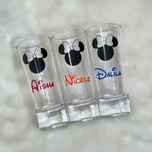 May include: Three clear shot glasses with a Minnie Mouse design. Each glass has a black silhouette of Minnie's head with a white bow. The names "Aisha" in red, "Nicole" in orange, and "Dalila" in blue are printed on the glasses.