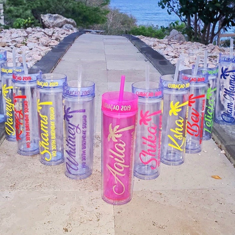 May include: A set of eight clear plastic tumblers with colorful names and palm tree designs. The tumblers are personalized with names like "Darry", "Shams", "Whitney", "Agila", "Shika", "Ksha", "Ber", and "Leticio". Each tumbler has a straw and the text "CURACAO 2019" on the side.