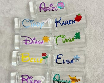 May include: Clear glass shot glasses with names like Ariel, Cindy, Karen, Diana, Tiana, Belle, Elsa, Riley, and Anna, each with a colorful design. The shot glasses are arranged on a white surface.