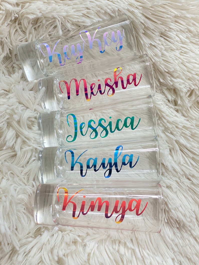 Puede incluir: Cinco vasos de chupito transparentes con nombres coloridos e iridiscentes. Los nombres son: Key Key, Meisha, Jessica, Kayla y Kimya.